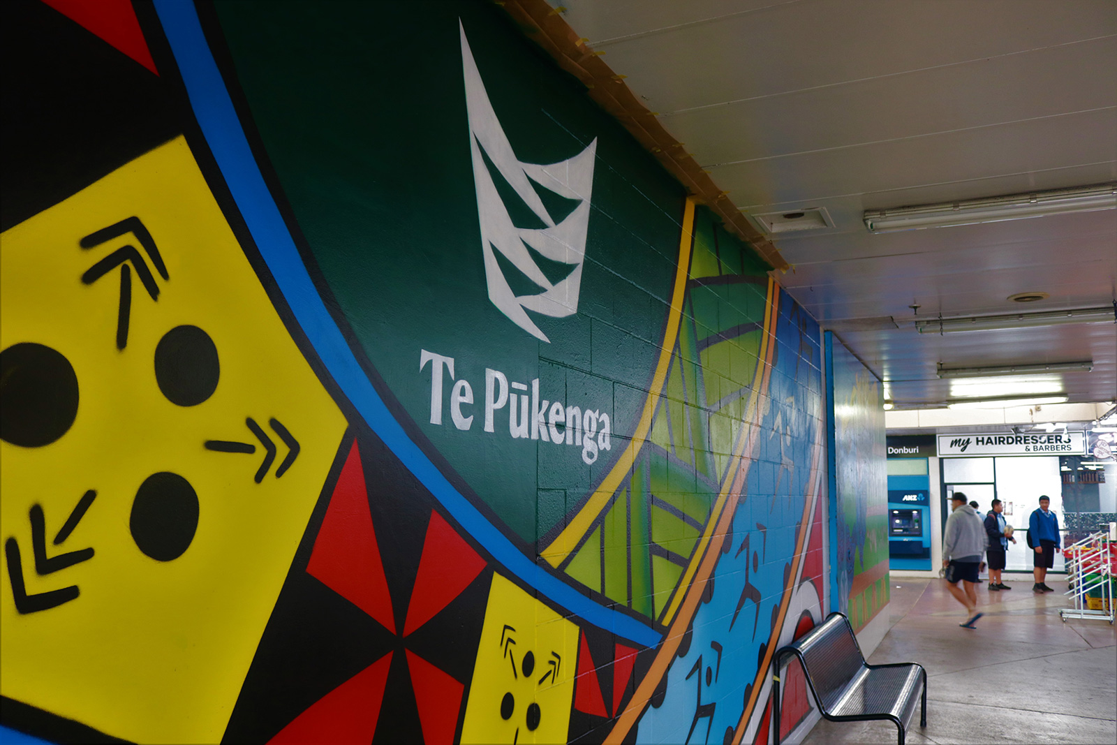 This image has been photographed from the left side of the mural. The mural contains multicultural patterns with Te Pūkenga logo at the top and centre of the mural. There is a metal seat up against the building that the mural is painted on and facing away. Looking to the right, in the background, there is an ANZ atm and a hairdressing shop.