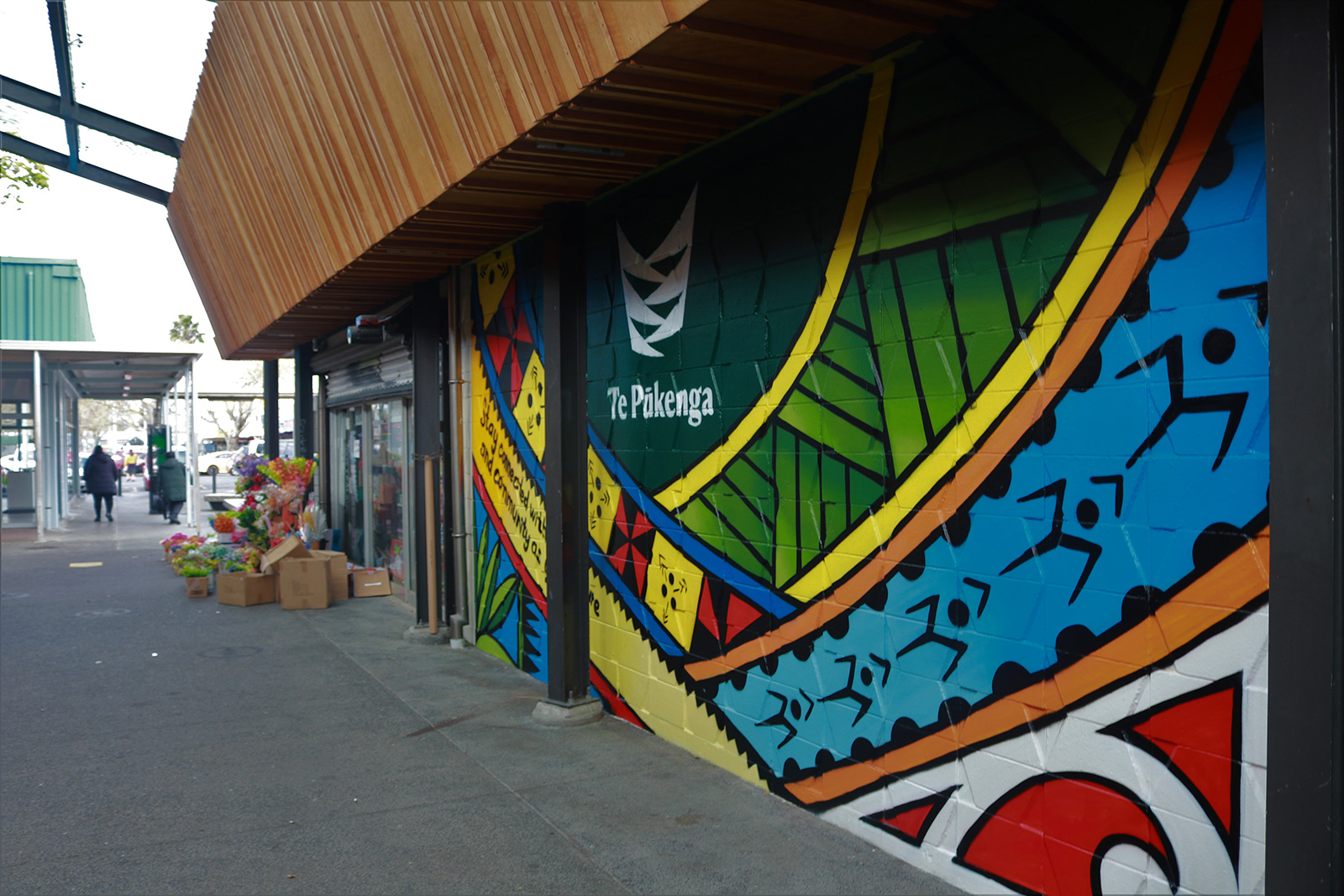 Image of a building that has a colourful mural depicting multicultural patterns with Te Pūkenga logo at the top and centre. The image has been taken to the right of the mural and in the distance there is a flower shop with flowers and cardboard boxed outside.
